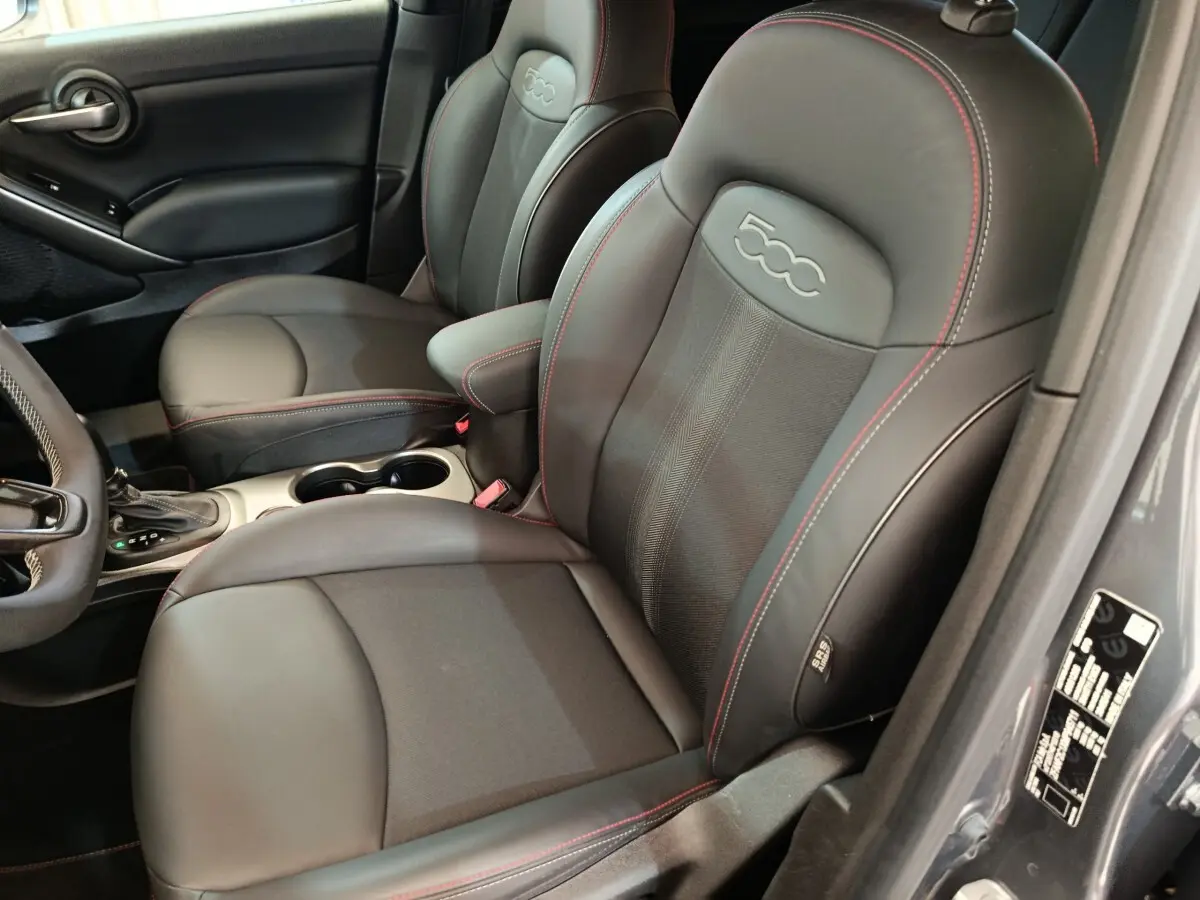 Vue intérieure côté conducteur du Fiat 500X 2021 gris foncé, sièges en cuir noir avec surpiqûres rouges et logo 500 embossé.