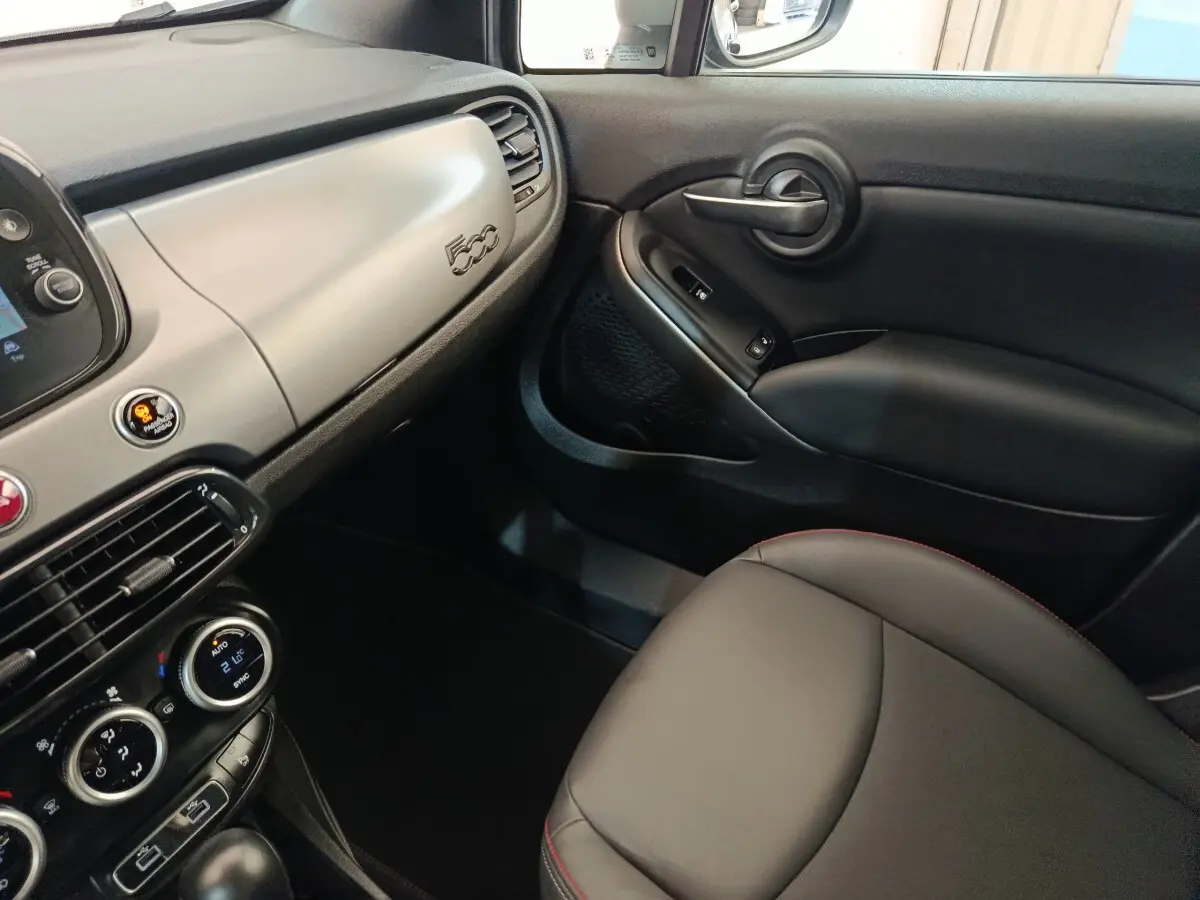 Intérieur côté passager de la Fiat 500X 2021, tableau de bord gris clair avec logo 500 et sellerie noire avec surpiqûres rouges.