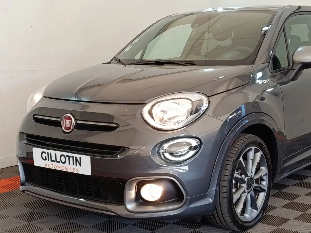 Vue 3/4 avant droite d'une FIAT 500X gris foncé avec phares allumés et badge Sport sur l'aile.