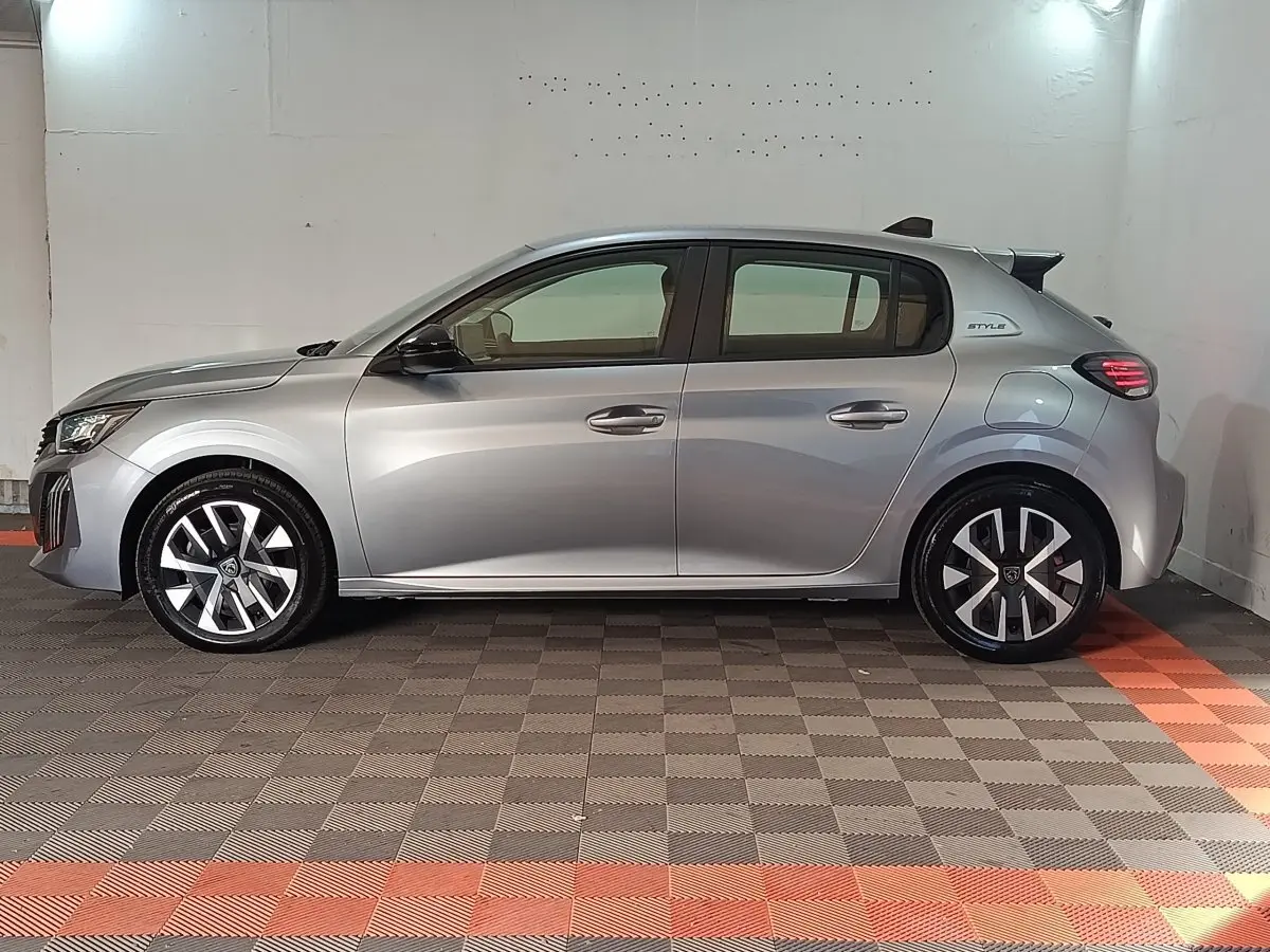 Profil droit de la Peugeot 208 Hybrid gris clair 2025 avec jantes stylisées et badge Style sur l'aile arrière.