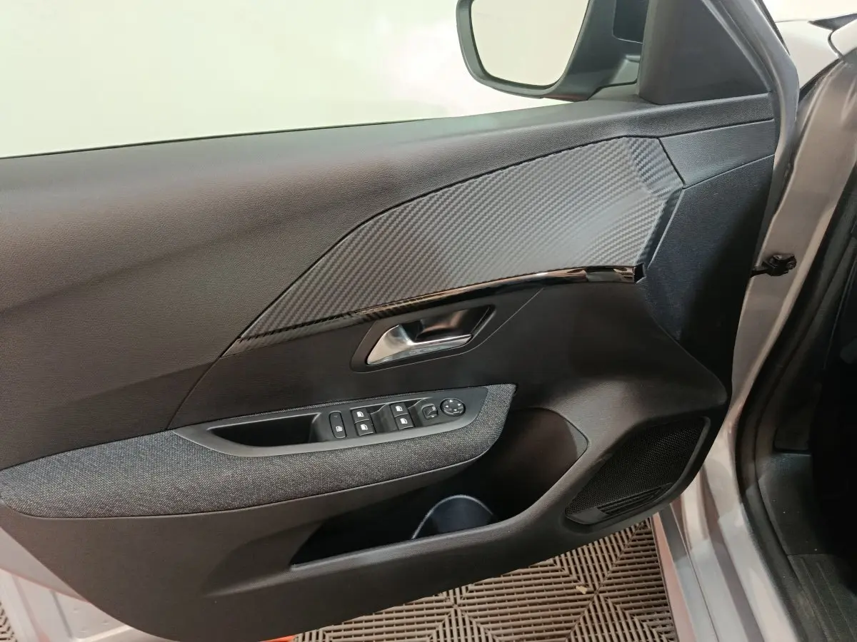 Vue intérieure du panneau de porte côté conducteur gris clair de la Peugeot 208 Hybrid 2025 avec commandes électriques et insert effet carbone.