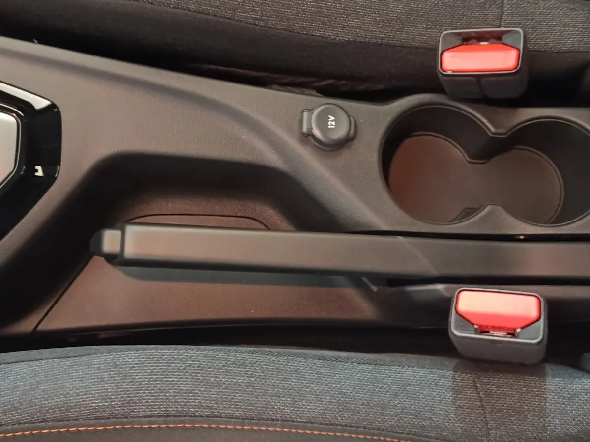 Gros plan sur la console centrale noire de la Peugeot 208 Hybrid 2025, montrant frein à main, prise 12V et porte-gobelets.
