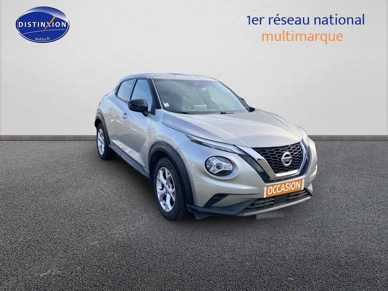 Nissan Juke gris clair vu de 3/4 avant droit avec calandre noire et feux ronds distinctifs.