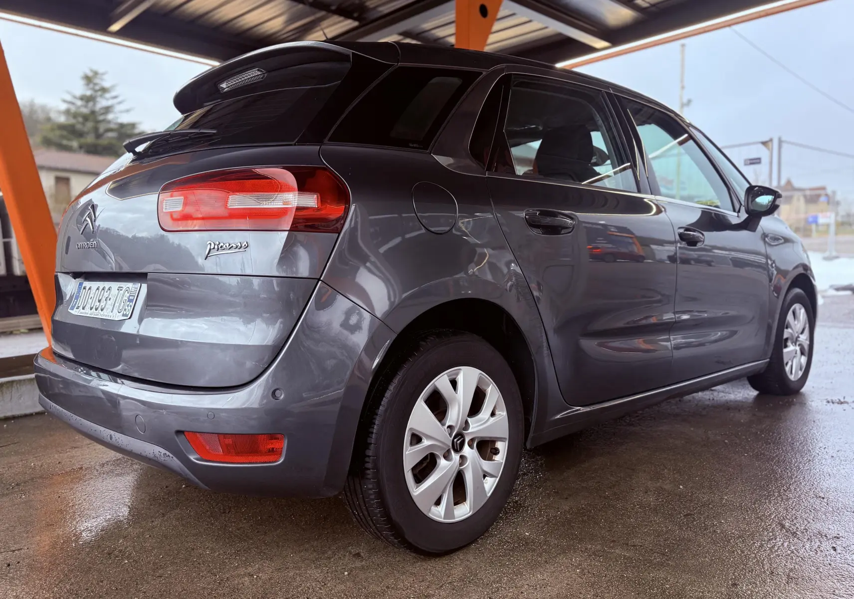 Vue 3/4 arrière droite d'une Citroën C4 Picasso gris platinium métallisé sous abri, avec feux arrière et jantes visibles.