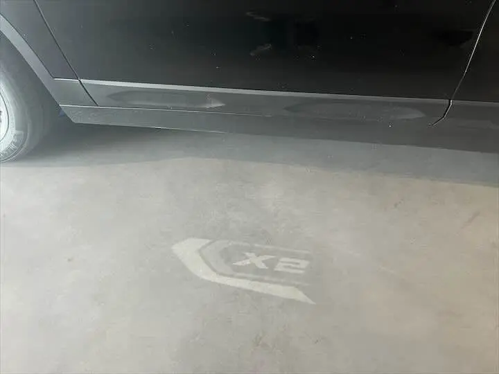 Projection lumineuse du logo BMW X2 au sol à côté de la porte côté gauche d'un SUV noir métallisé.