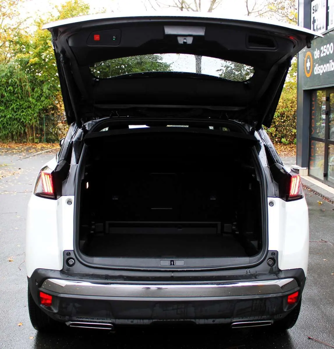 Vue arrière du Peugeot 3008 blanc nacré 2025 avec coffre ouvert et feux arrière allumés sur parking extérieur humide.
