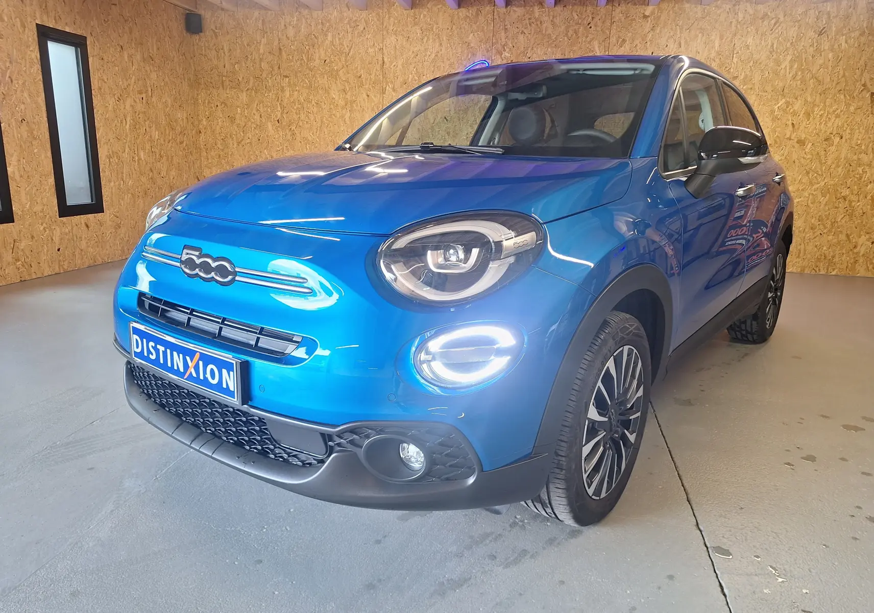 Fiat 500X bleu métallisé vue 3/4 avant droit avec phares LED et jantes alliage noires dans un garage.