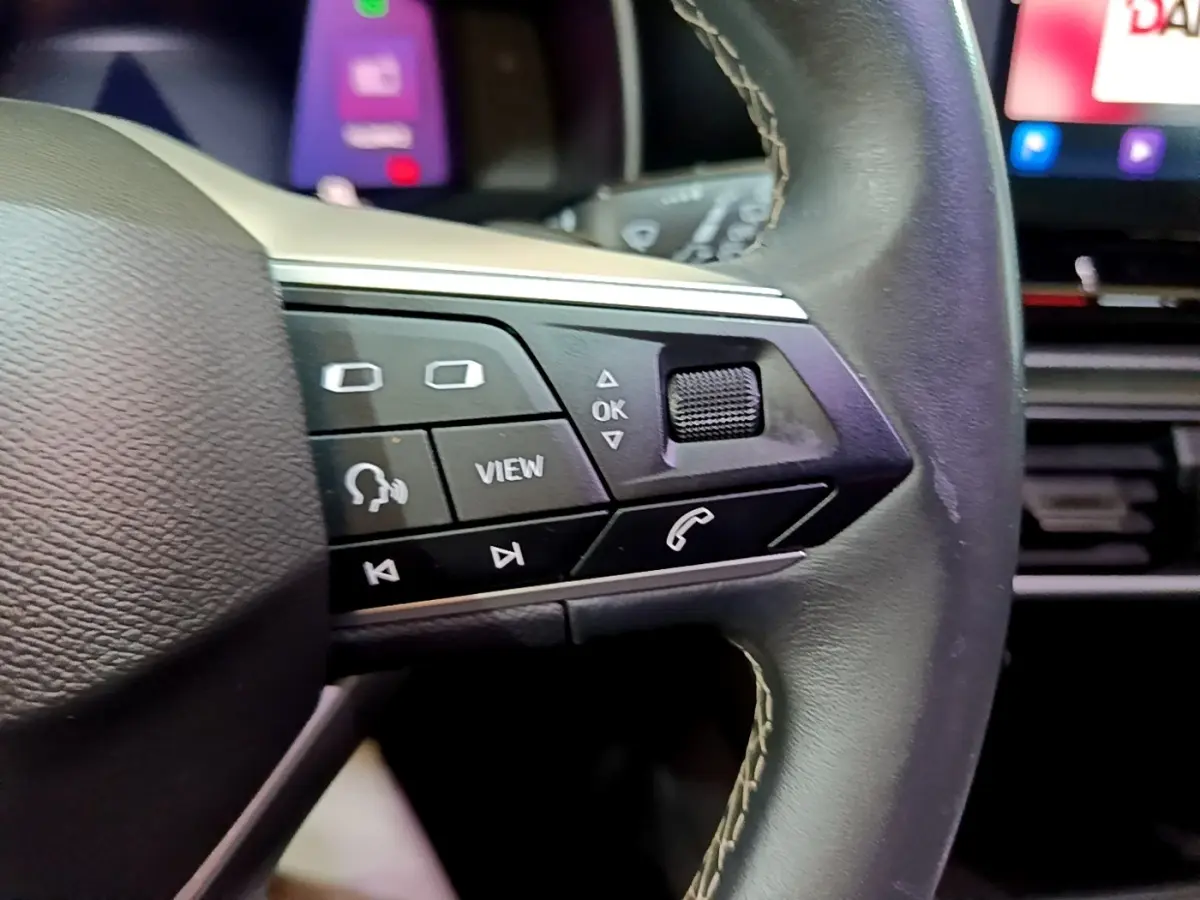 Gros plan sur les commandes à droite du volant cuir noir du SEAT Leon gris foncé, avec écran tactile en arrière-plan flou.