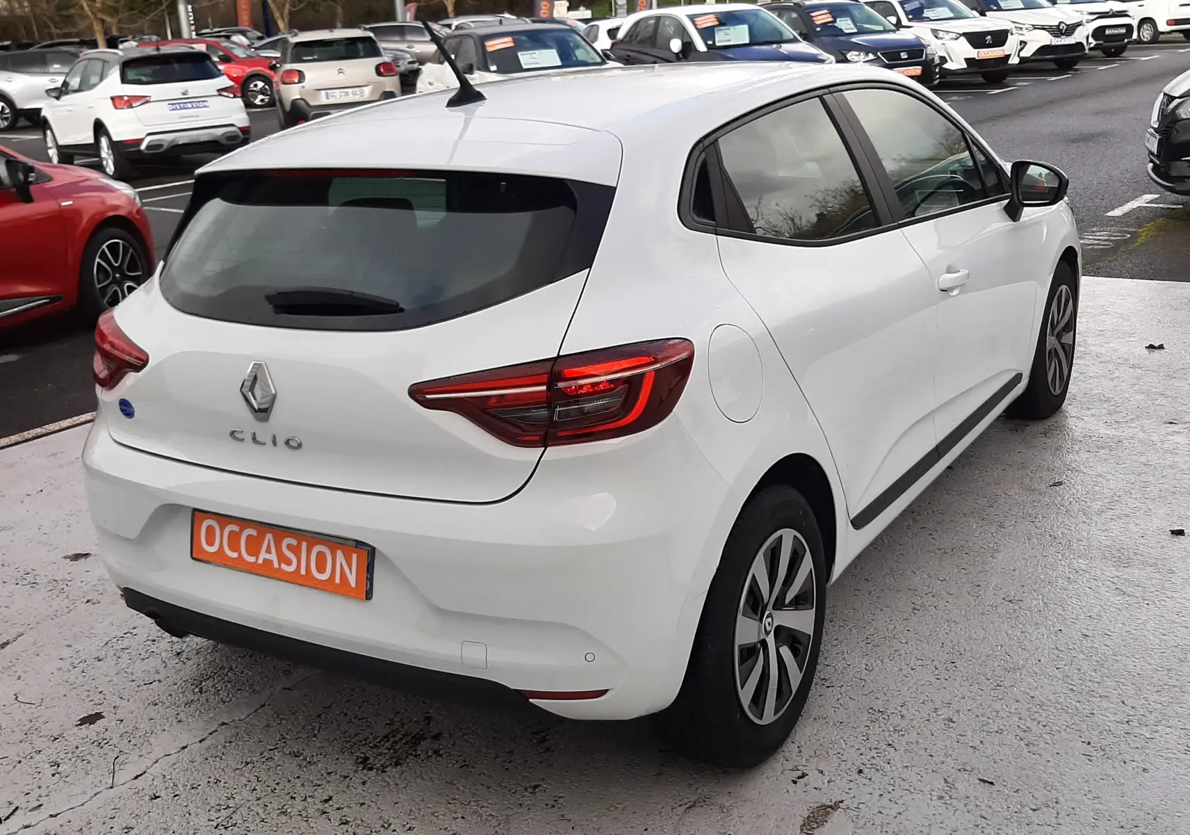 Vue 3/4 arrière droite d'une Renault Clio V blanche 5 portes avec feux LED et plaque "OCCASION" sur parking.