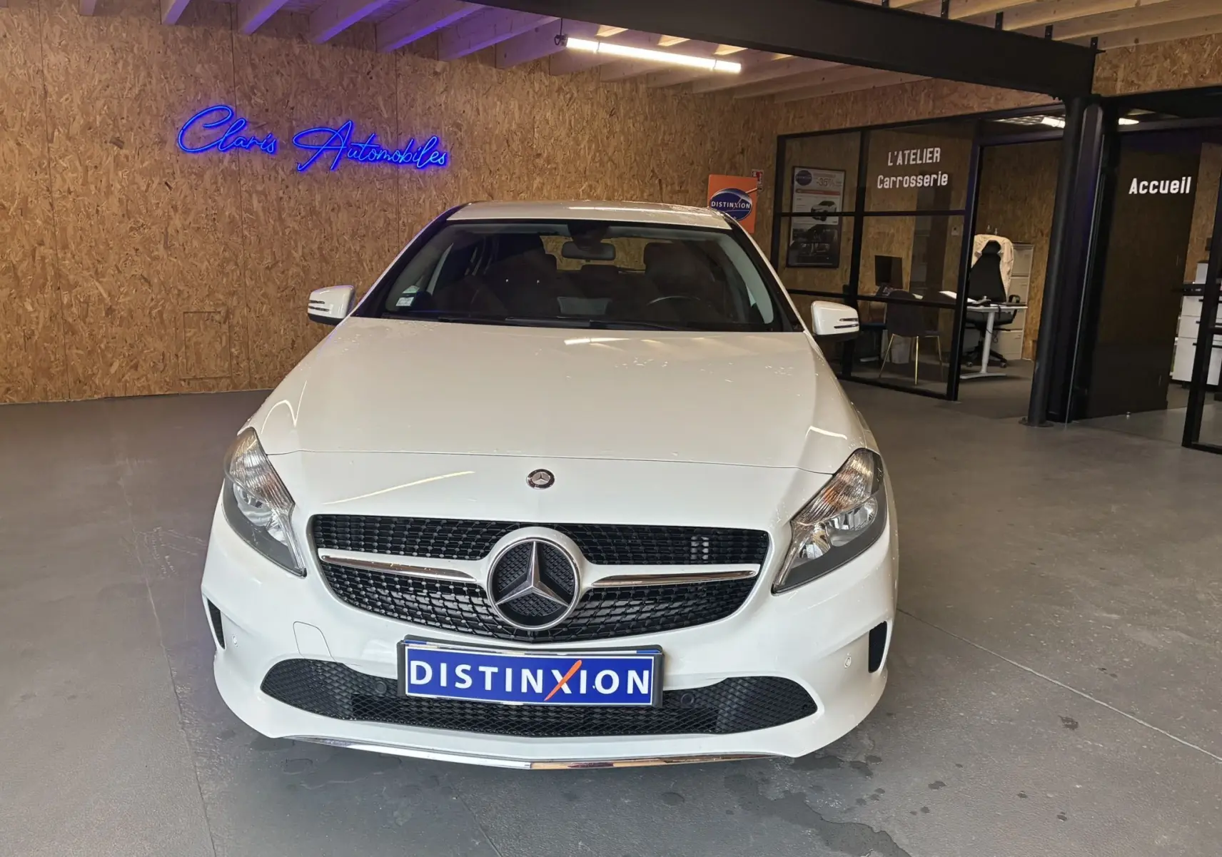 Vue frontale d'une Mercedes Classe A 200D blanche dans un showroom avec un mur en bois et enseigne néon bleue.