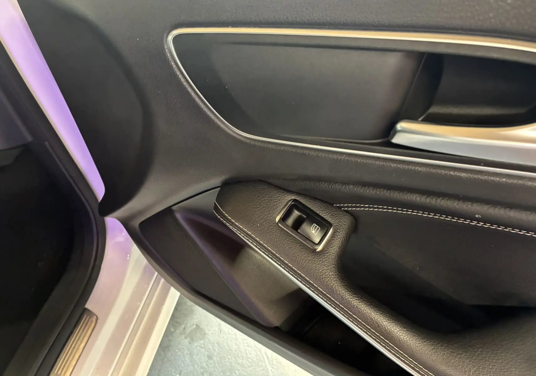Gros plan sur la poignée intérieure côté gauche d'une Mercedes Classe A blanche, avec bouton de commande de vitre et surpiqûres blanches.