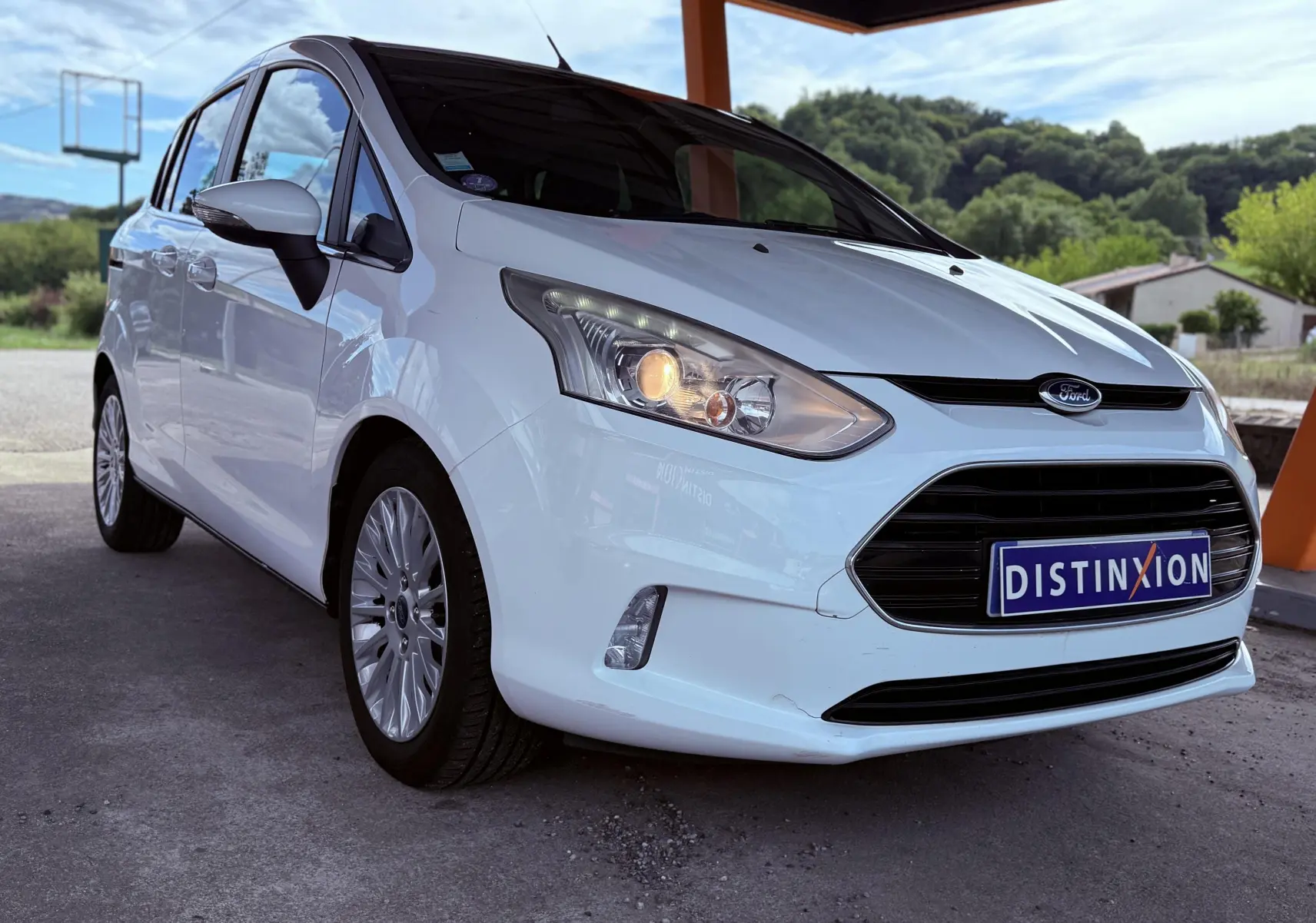 Ford B-MAX blanc vu en 3/4 avant droit sous un abri, avec phares allumés et jantes alliage.