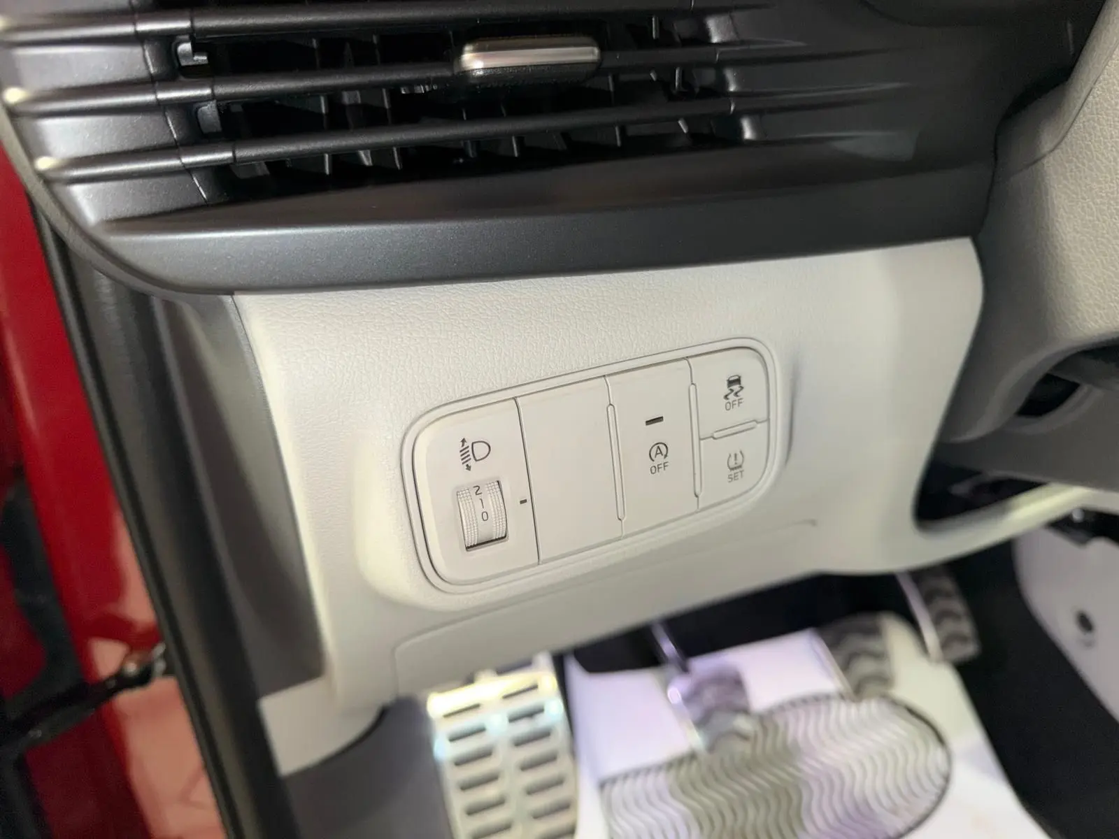 Gros plan sur les commandes de réglage des feux et aides à la conduite, avec pédales sport et intérieur bi-ton gris du Hyundai Bayon rouge.
