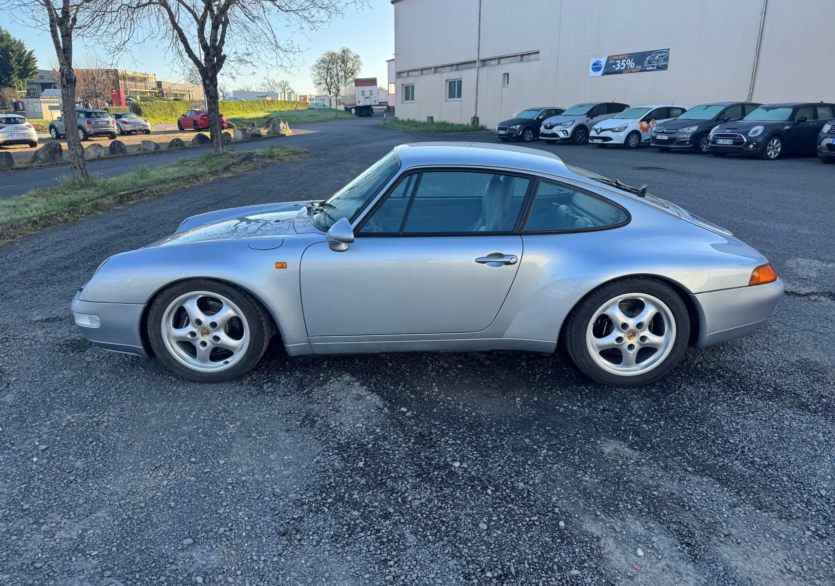 Profil côté gauche d'une Porsche 911 Carrera 2 type 993 gris polaire, avec jantes alliage et pare-brise teinté dégradé.