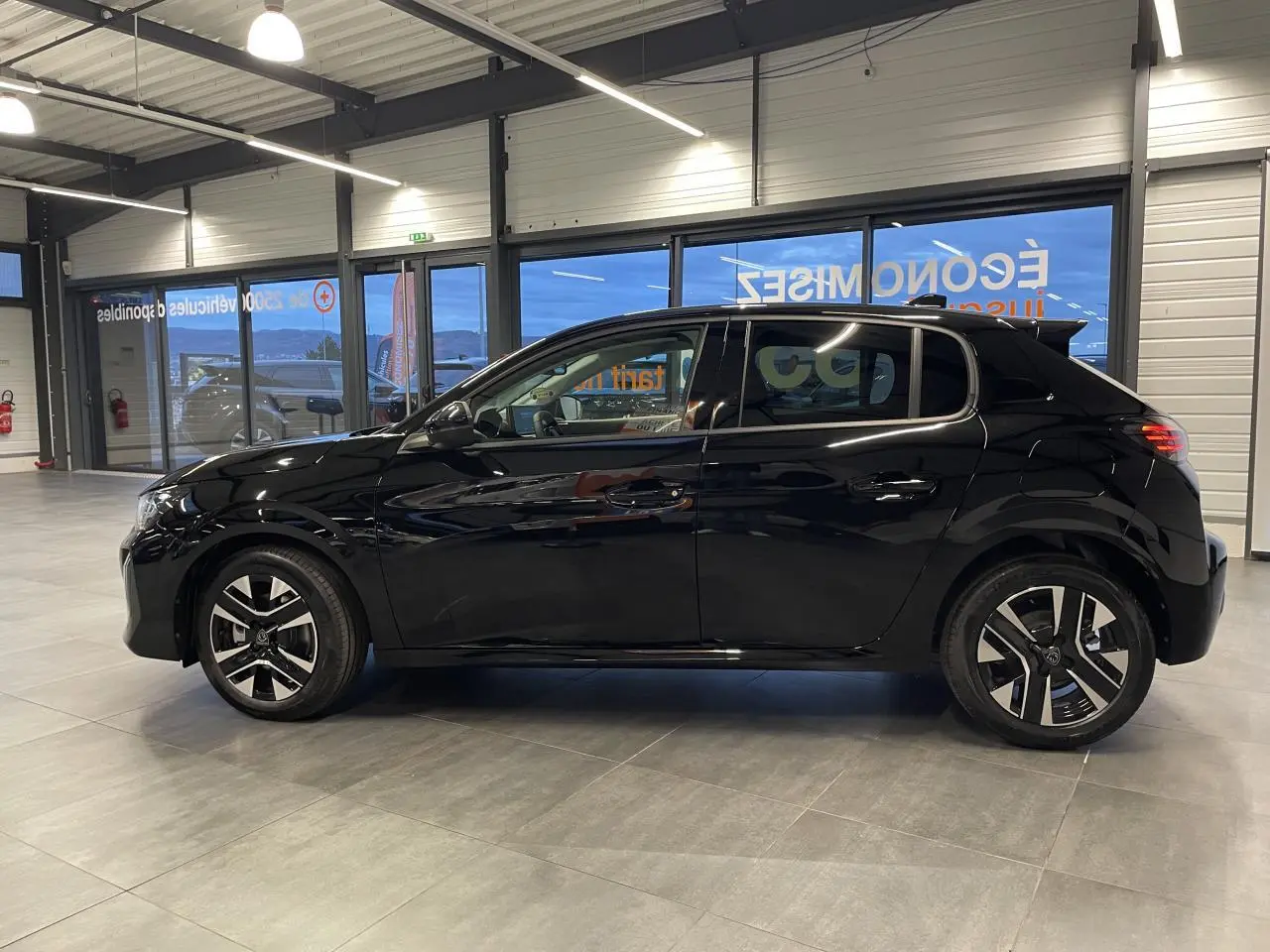 Profil droit d'une Peugeot 208 noire métallisée 2025 en showroom, avec jantes alliage bi-tons et vitres teintées.