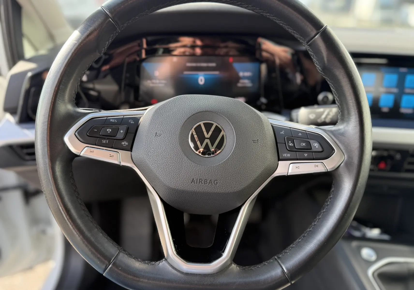 Vue rapprochée du volant en cuir noir avec logo Volkswagen, tableau de bord digital et écran tactile en arrière-plan d'une Golf blanche.