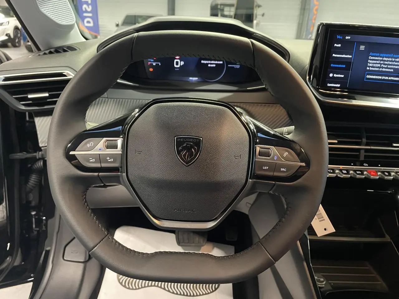 Vue rapprochée du volant cuir noir de la Peugeot 208 1.2i Hybrid 2025 avec tableau de bord numérique et écran tactile.