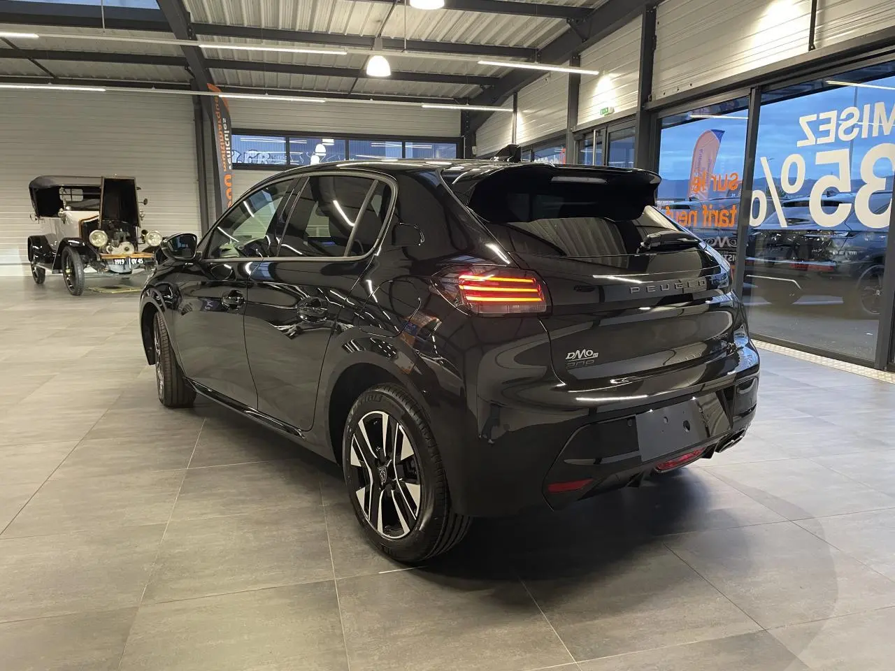 Vue 3/4 arrière droite d'une Peugeot 208 noire métallisée avec feux arrière allumés en showroom.
