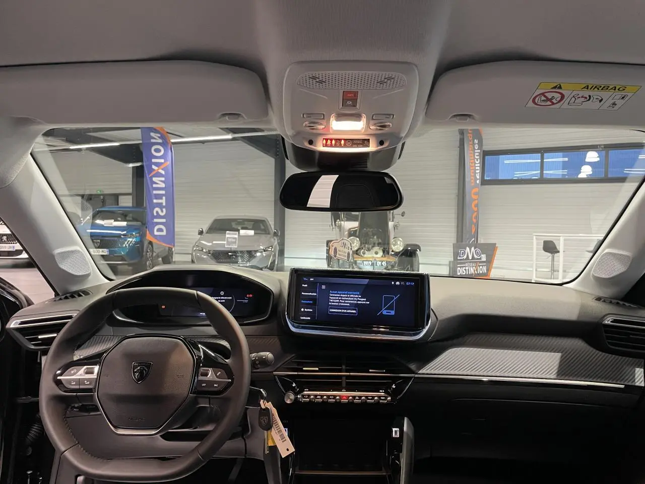 Vue intérieure avant du tableau de bord noir et volant compact cuir de la Peugeot 208 1.2i Hybrid 2025 avec écran tactile central.