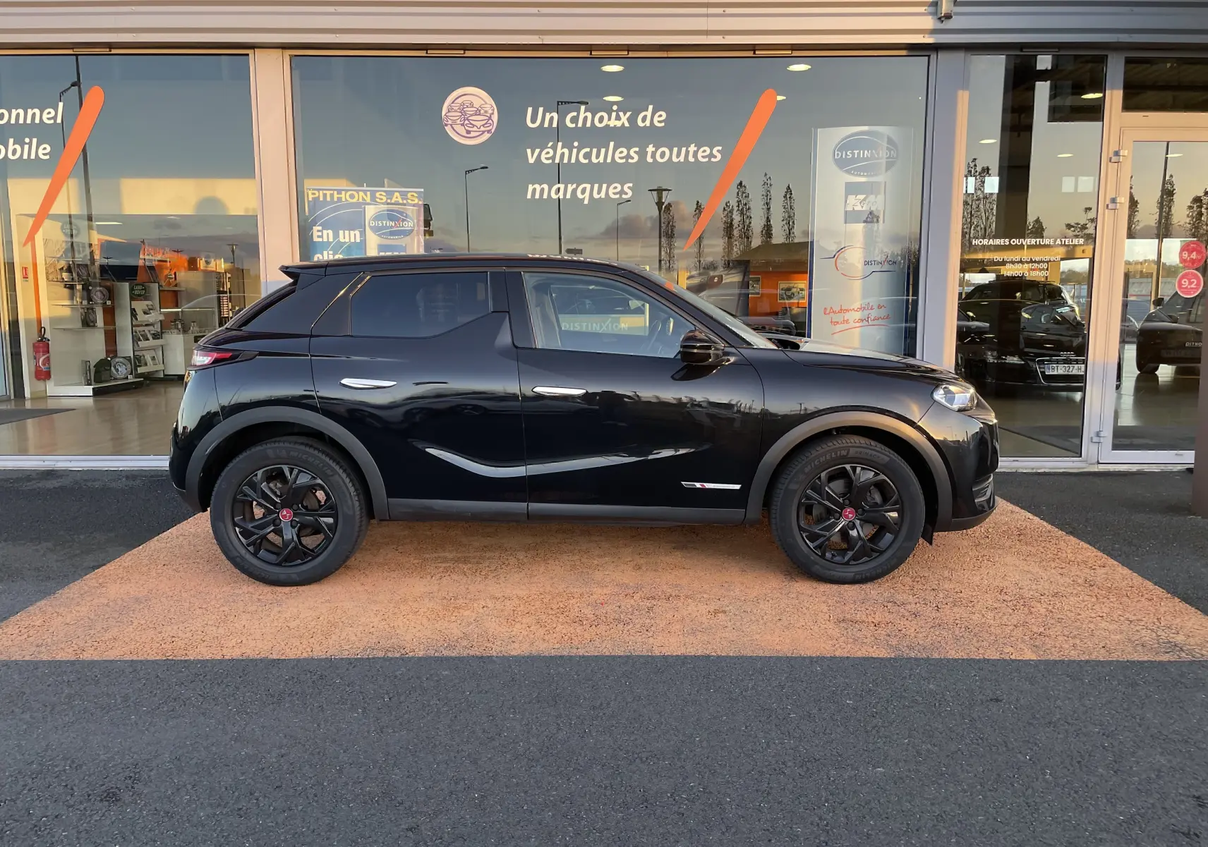 Profil droit d'un DS3 Crossback noir métal avec jantes noires, garé devant une vitrine de concession.