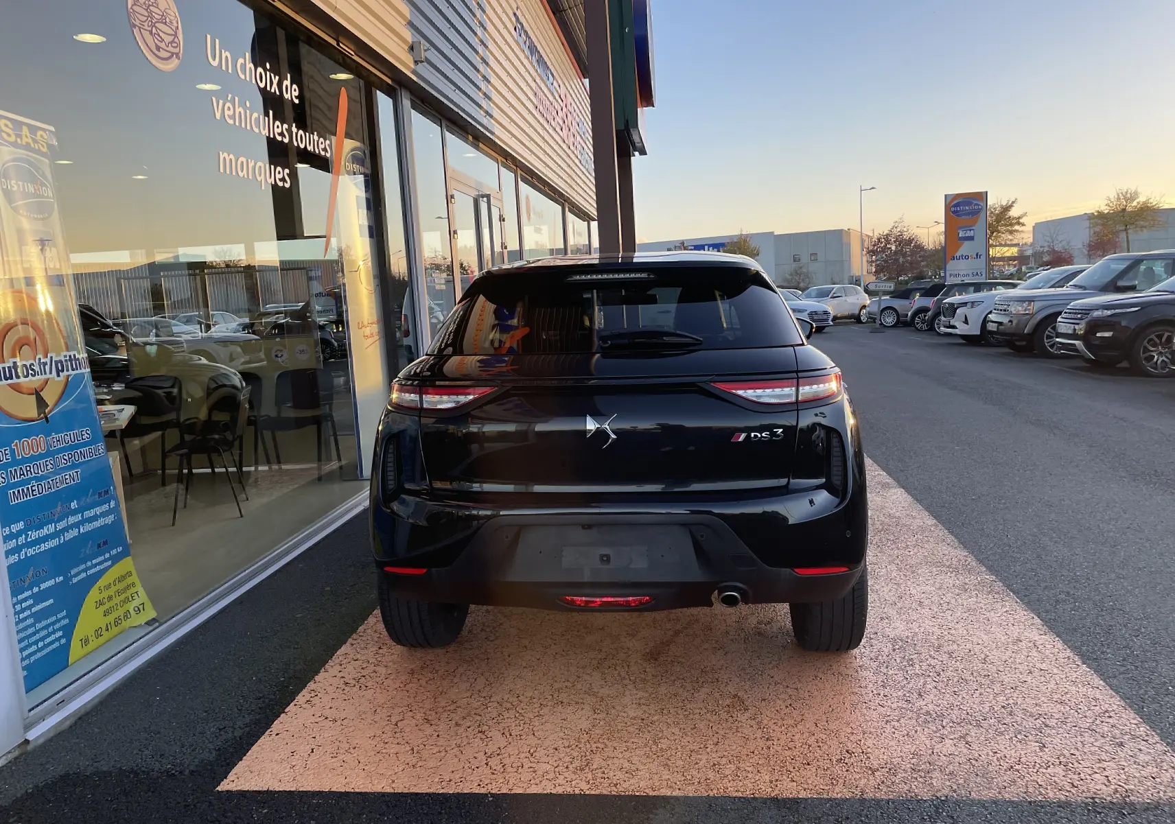 Vue arrière d'un DS3 Crossback noir métal garé devant un showroom, avec feux arrière allumés au crépuscule.
