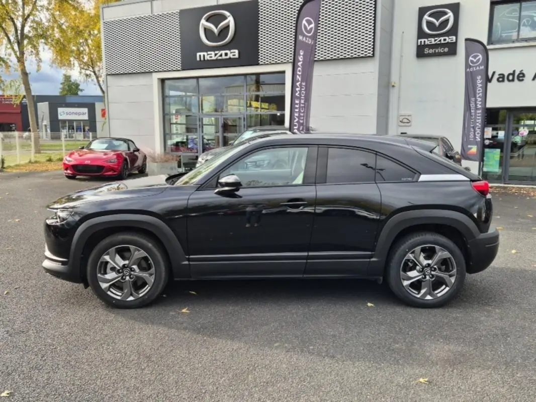 Vue de profil côté gauche d'un Mazda MX-30 électrique noir devant un concessionnaire Mazda avec logos visibles.