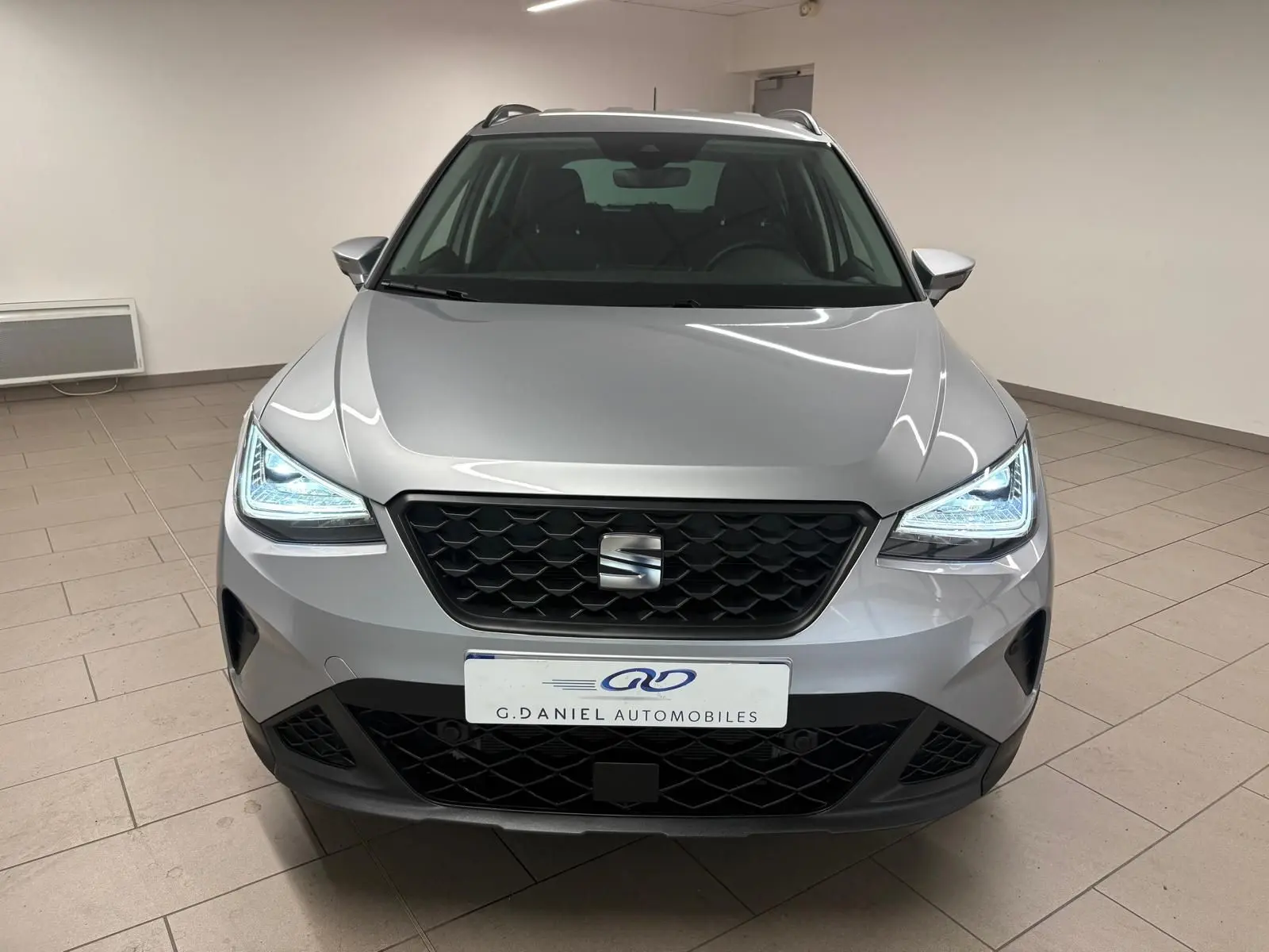 Vue frontale d'un SEAT Arona 2024 gris urbain avec phares LED allumés et calandre noire en intérieur showroom.