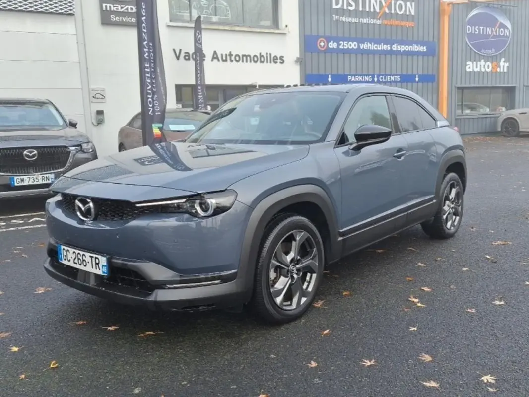 Vue 3/4 avant d'un Mazda MX-30 gris électrique, avec jantes distinctives et protections noires autour des passages de roues.