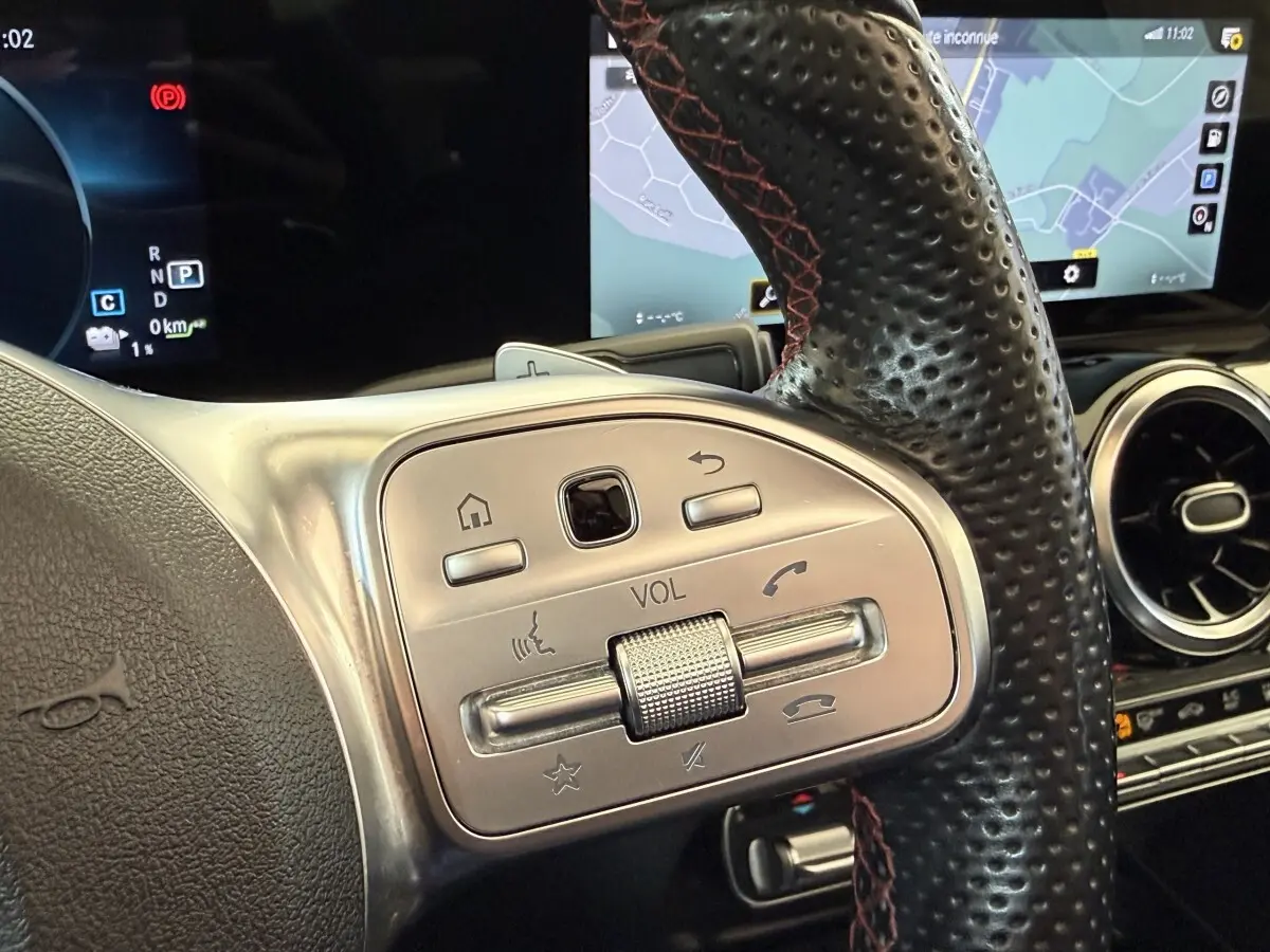 Gros plan sur les commandes argentées du volant en cuir noir perforé du Mercedes GLA 250 e AMG Line 2023, avec écran tactile en arrière-plan.