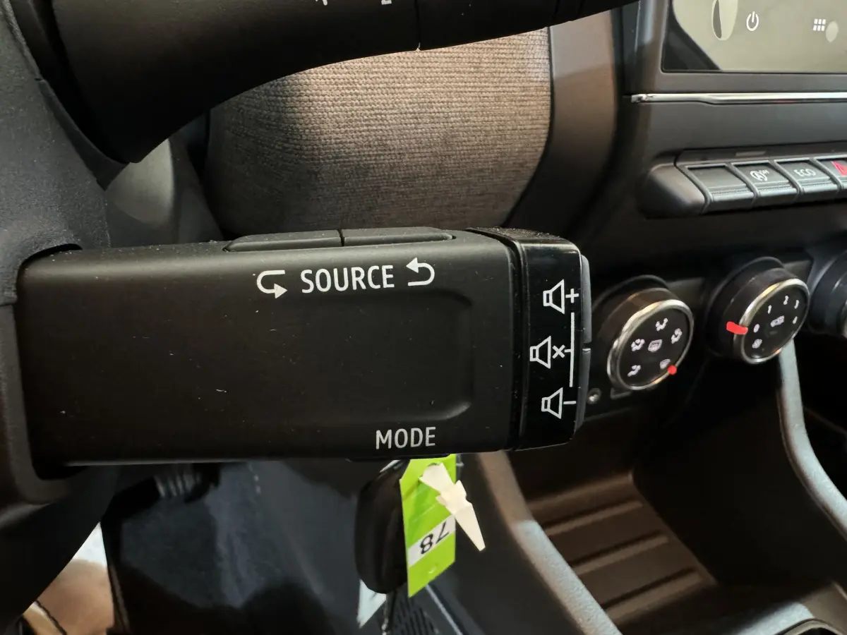 Gros plan sur la commande audio au volant d’une Renault Clio gris clair, avec tableau de bord et commandes de climatisation en arrière-plan.