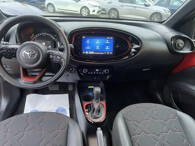 Vue intérieure avant de la Toyota Aygo X 2023, tableau de bord noir avec touches rouges et écran tactile central.