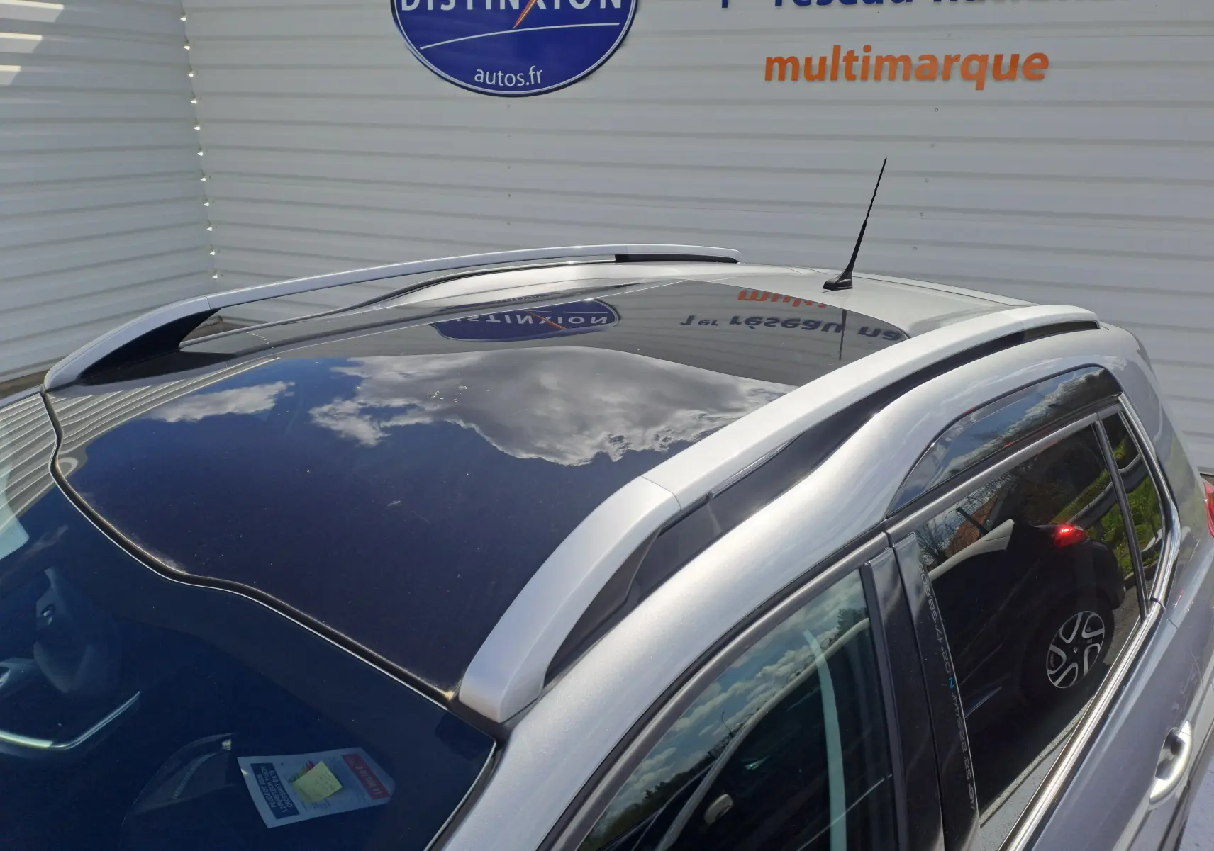 Vue en plongée du toit panoramique noir et des barres de toit argentées d'un Peugeot 2008 gris stationné près d'un mur blanc.