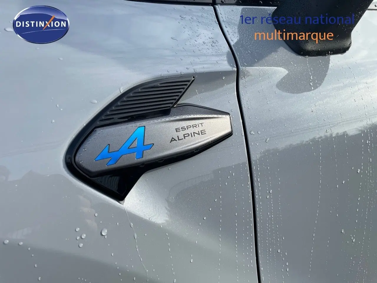 Gros plan sur l'écusson Esprit Alpine bleu sur la portière côté gauche d'une Renault Symbioz gris Rafale mouillée.