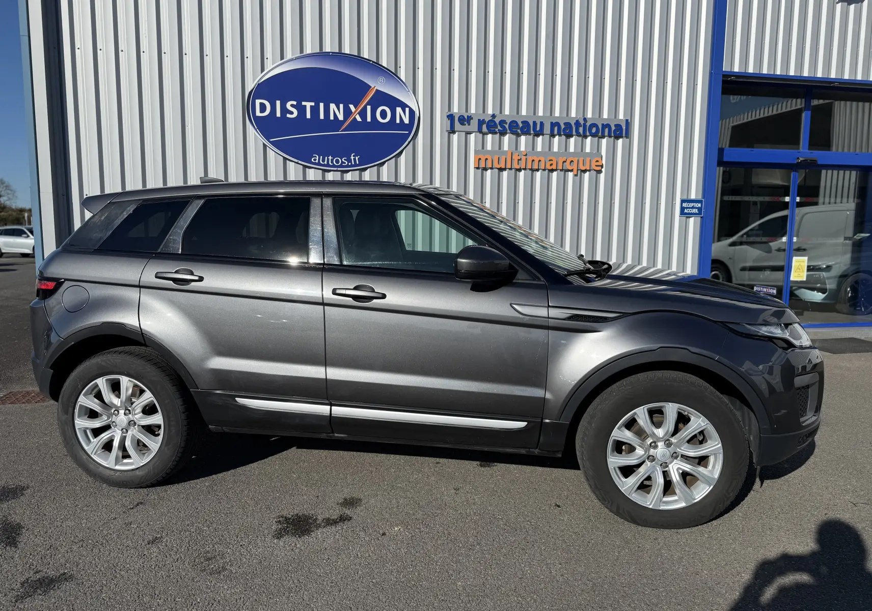 Vue de profil côté gauche d'un Range Rover Evoque gris métallisé garé devant un bâtiment avec enseigne Distinxion.