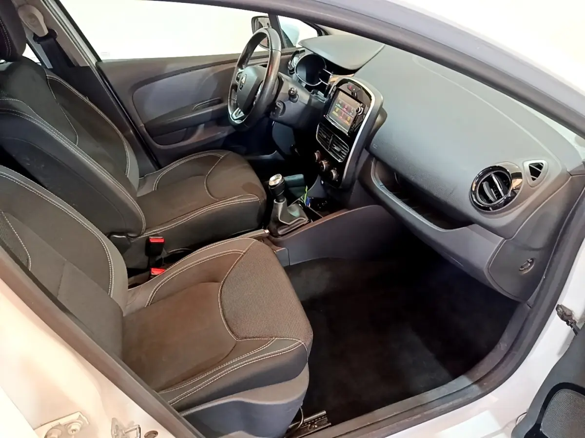 Vue intérieure côté conducteur de la Renault Clio blanche 2016, montrant sièges noirs et tableau de bord avec écran tactile.