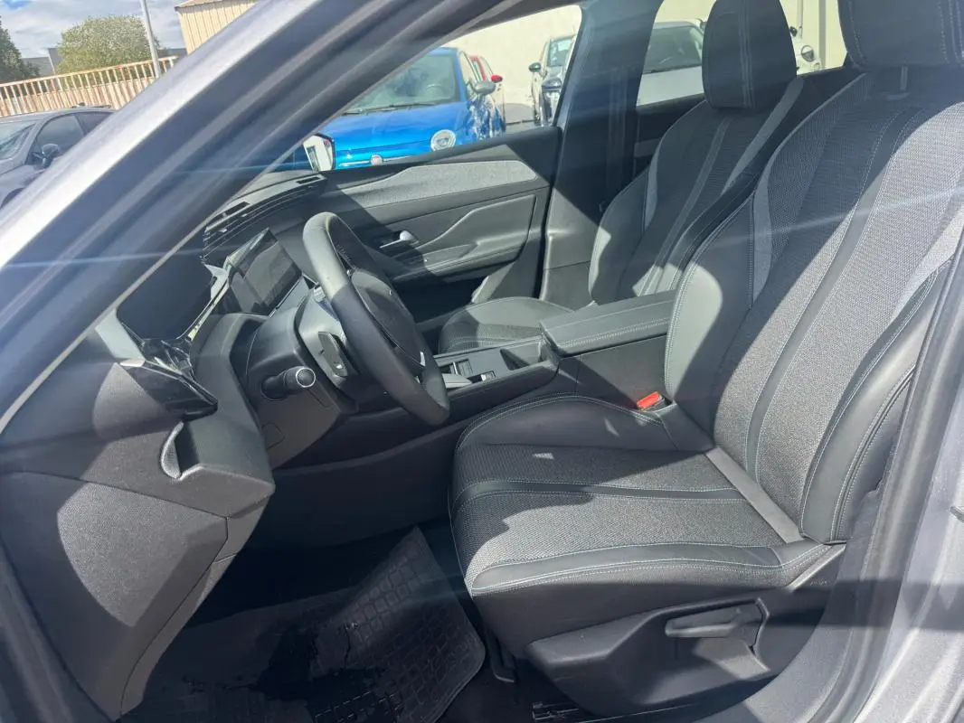 Vue intérieure côté conducteur de la Peugeot 308 Hybrid 145 gris, avec sièges tissu et accoudoir central visible.
