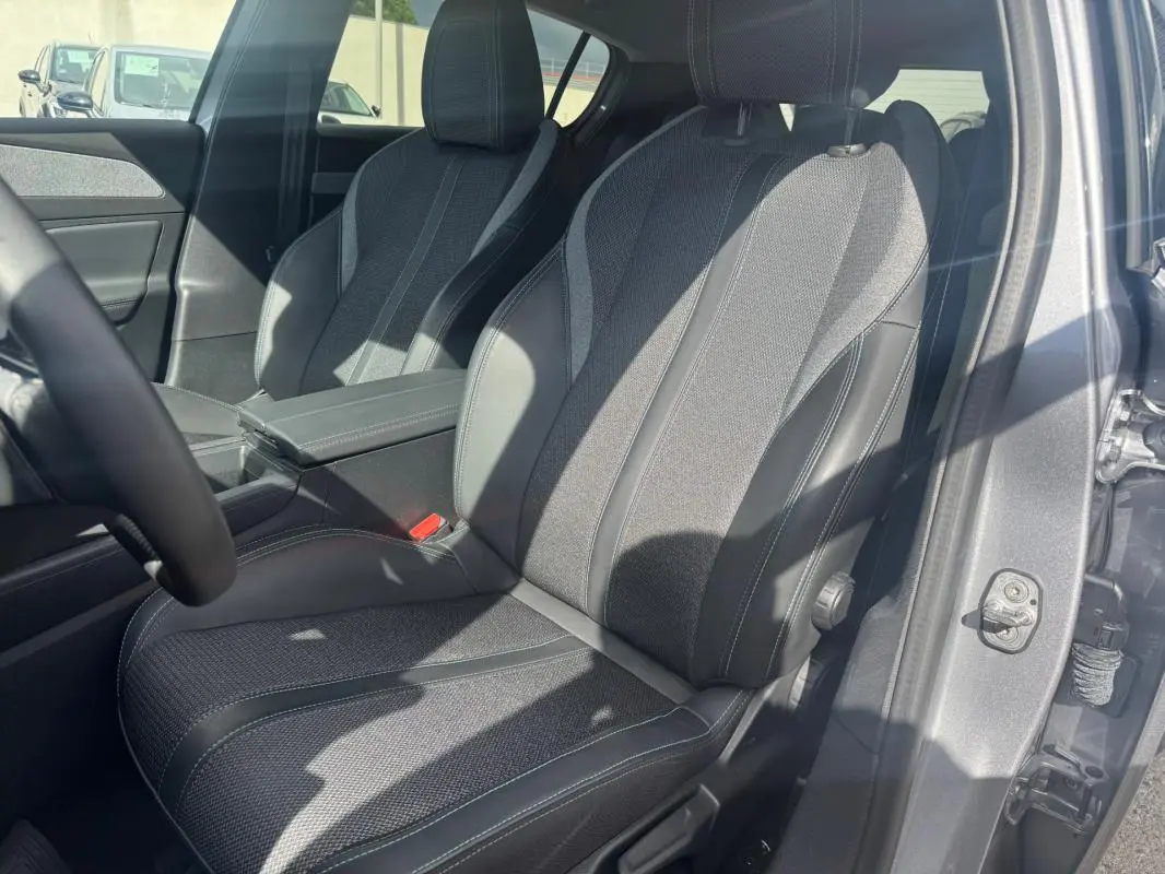 Vue intérieure côté conducteur d’un Peugeot 308 gris 2025, sièges tissu et cuir avec accoudoir central visible.