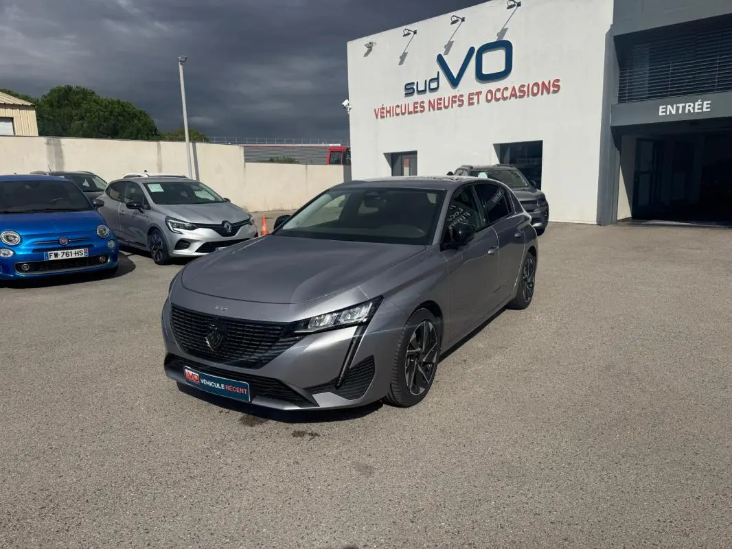 Peugeot 308 Hybrid gris vue 3/4 avant droit, jantes alliage 17 pouces, stationnée devant un bâtiment de vente auto.