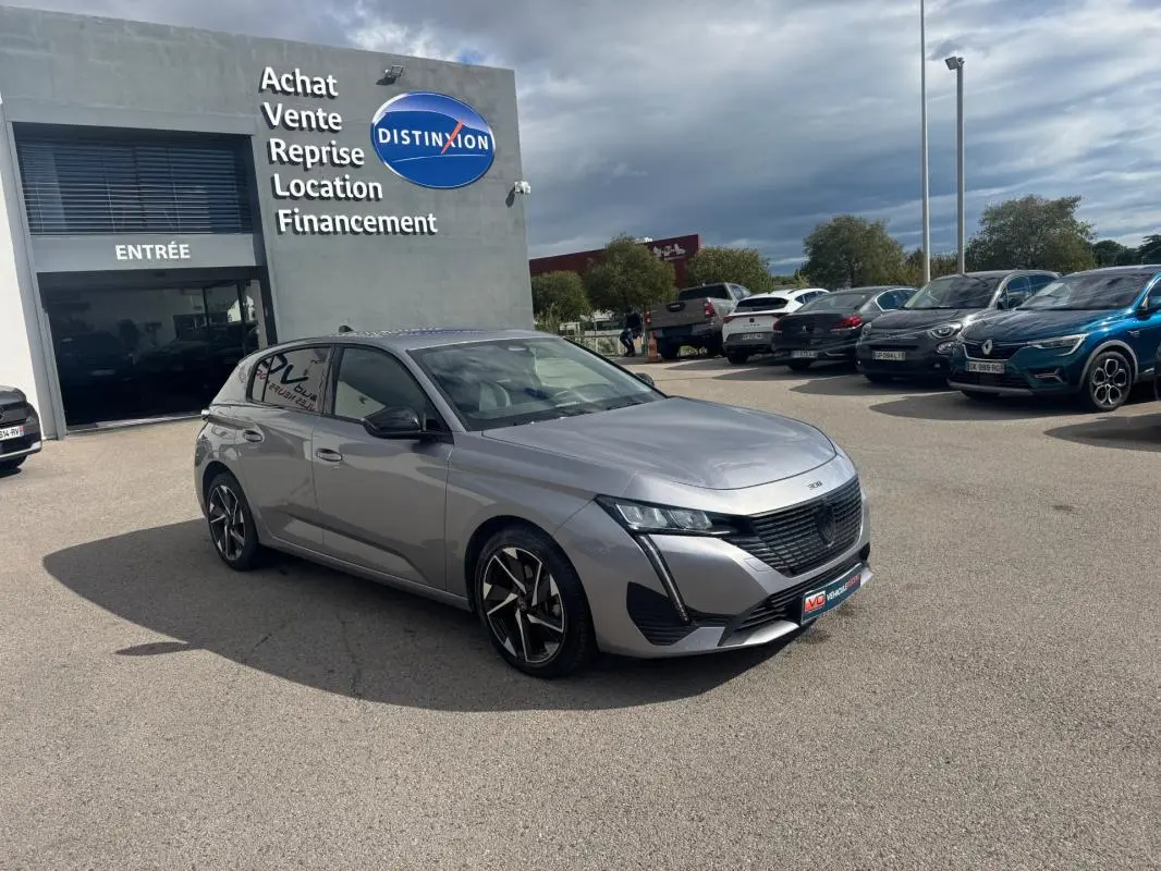 Peugeot 308 Hybrid gris vue 3/4 avant droit, avec jantes alliage 17 pouces, stationnée devant un concessionnaire.
