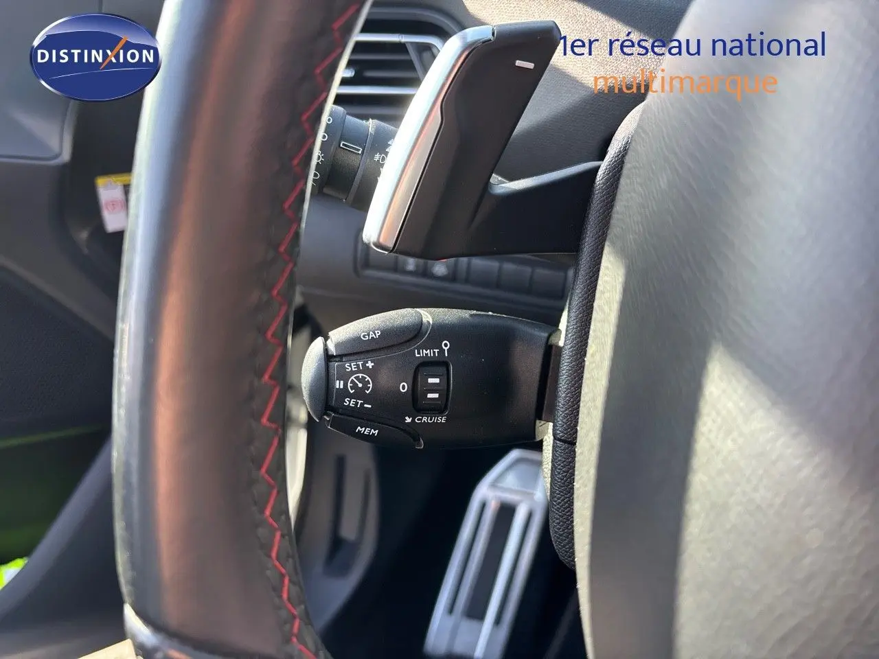 Gros plan sur la commande de régulateur de vitesse derrière le volant cuir noir avec surpiqûres rouges d'une Peugeot 308 blanche nacrée.