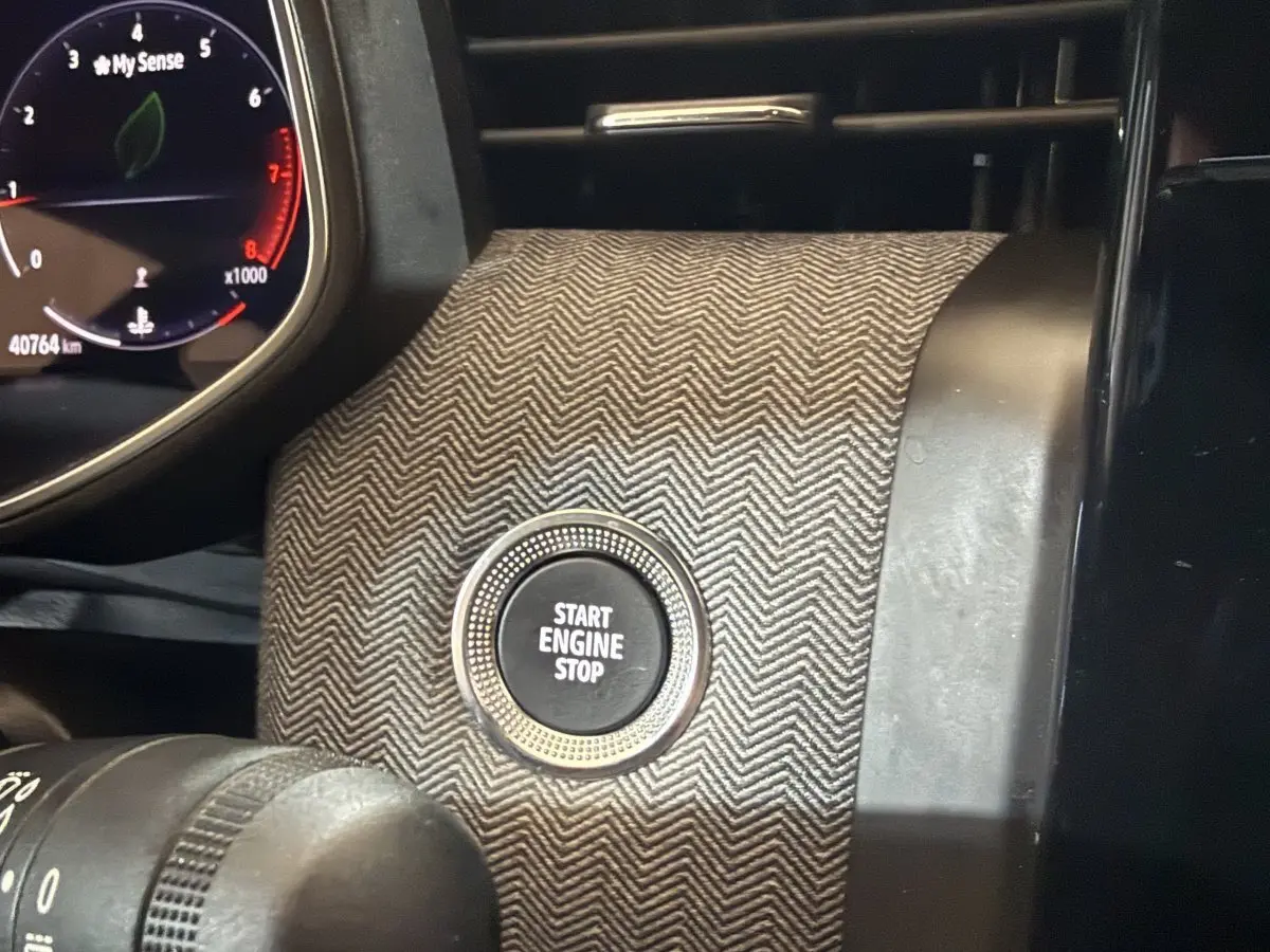 Gros plan sur le bouton Start Engine Stop entouré d’un habillage gris à motif chevrons, avec le combiné d’instruments partiellement visible.