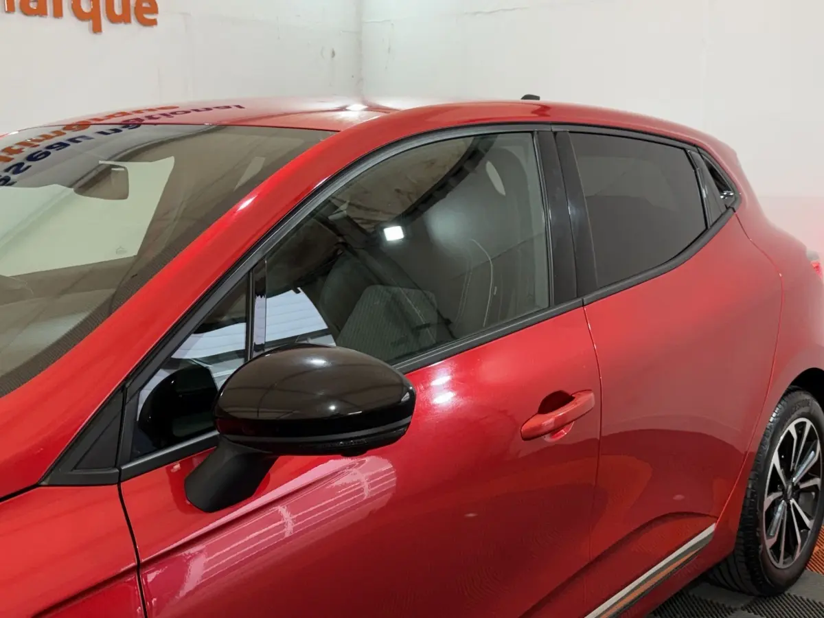 Vue latérale gauche rapprochée d’une Renault Clio rouge 2024 avec rétroviseur noir et vitres teintées.
