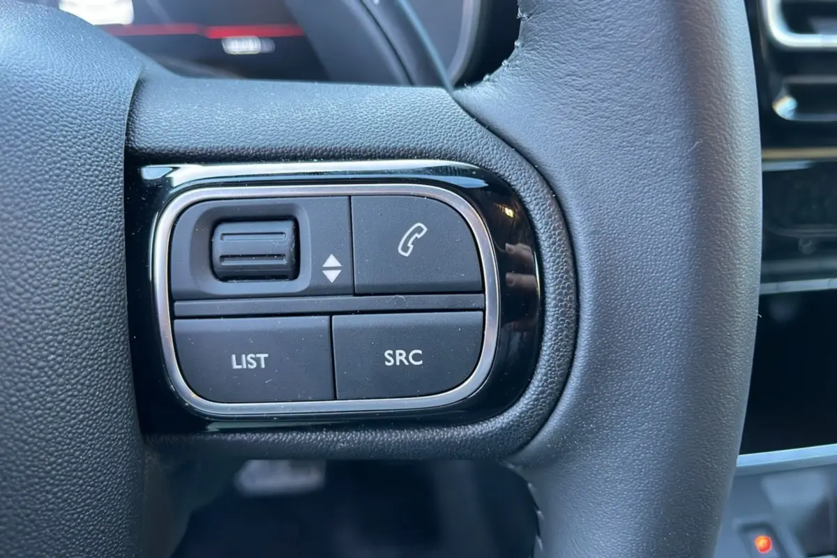 Gros plan sur les commandes multifonctions noires à gauche du volant cuir d’un Citroën C5 Aircross Hybrid blanc.