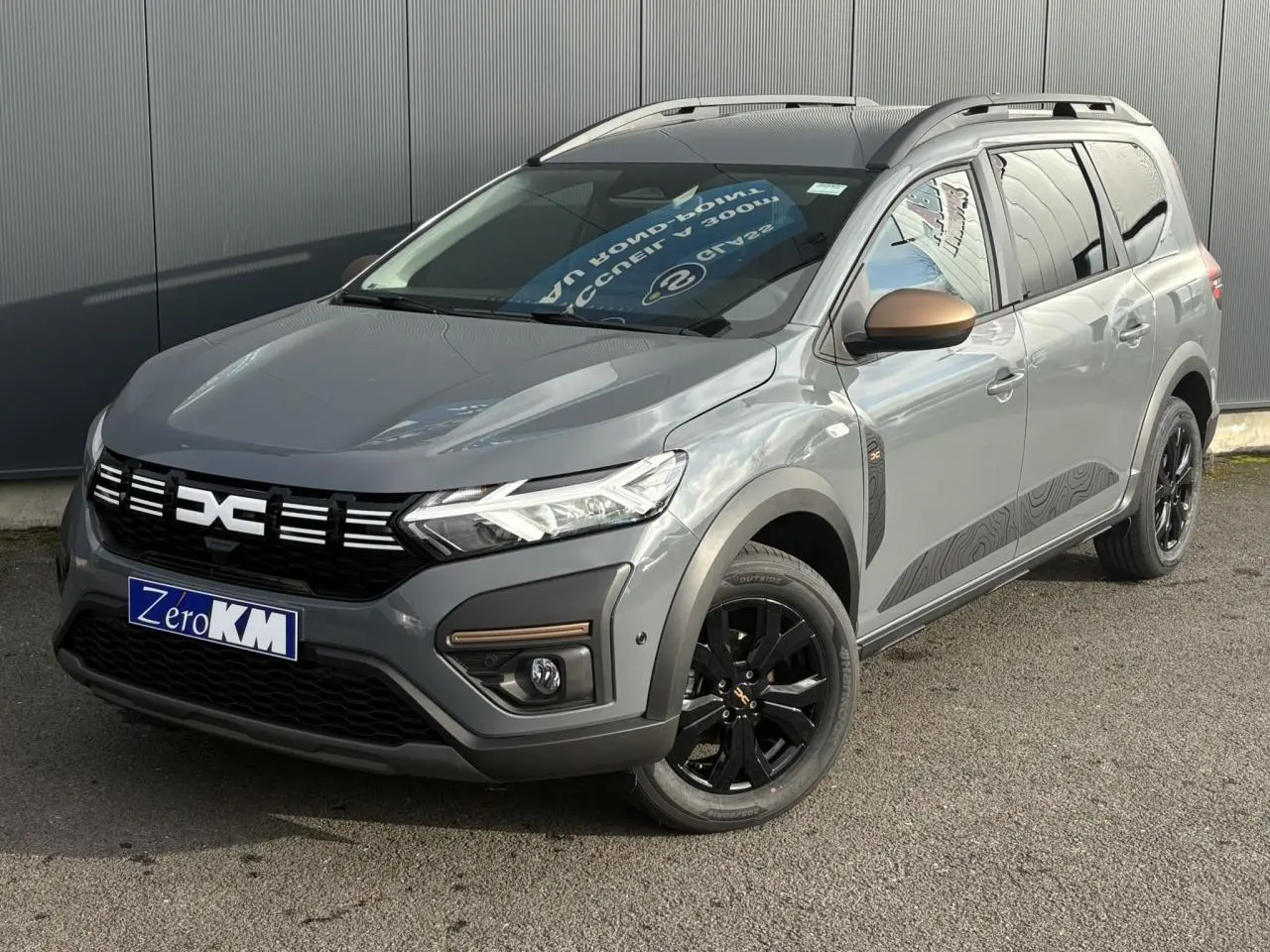 Dacia Jogger Gris Urbain en 3/4 avant droit avec jantes noires et barres de toit modulaires visibles.