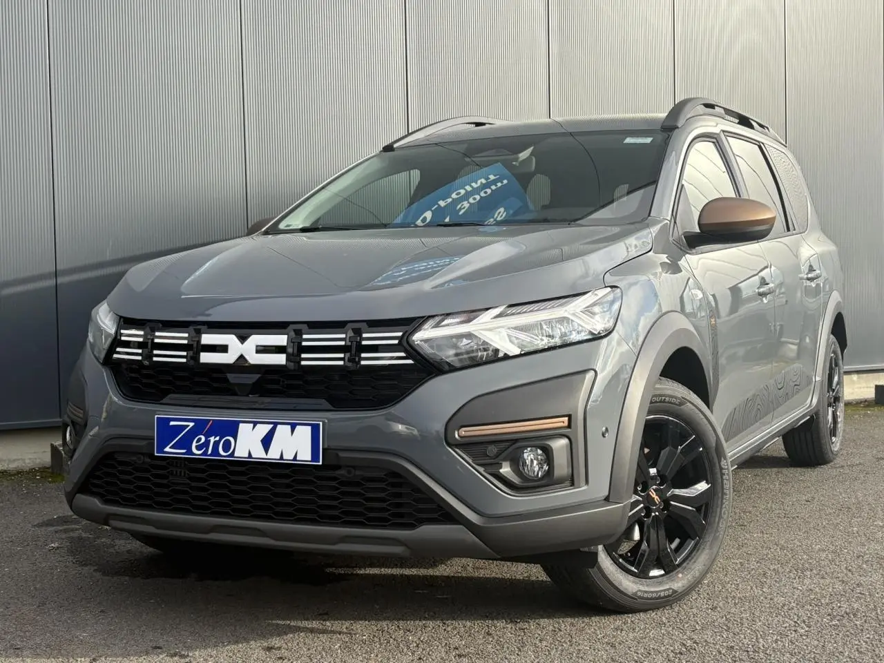 Dacia Jogger Gris Urbain en 3/4 avant droit avec jantes noires et barres de toit modulaires visibles.
