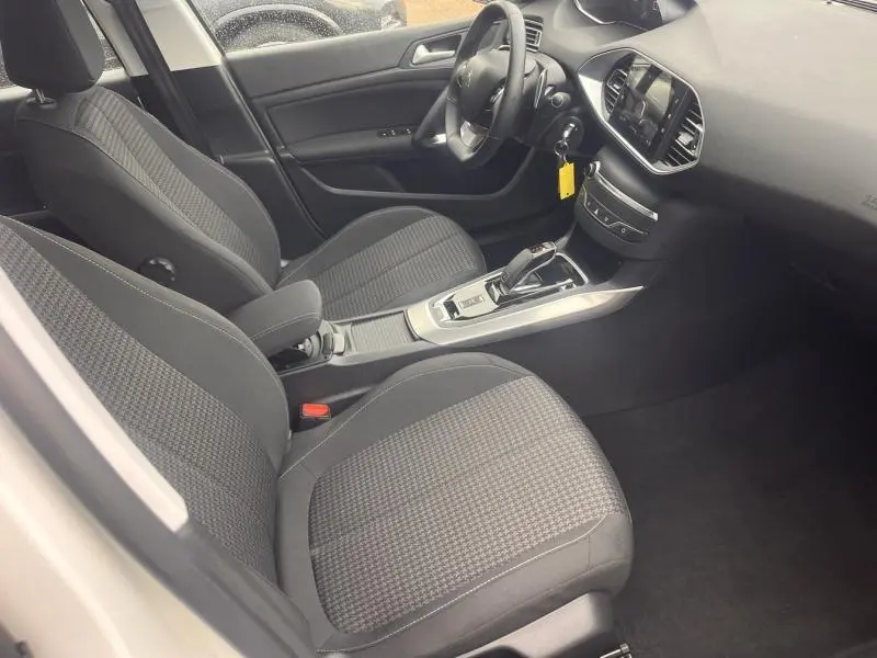 Vue intérieure côté conducteur de la Peugeot 308 blanche, montrant sièges tissu gris et console centrale avec boîte auto.