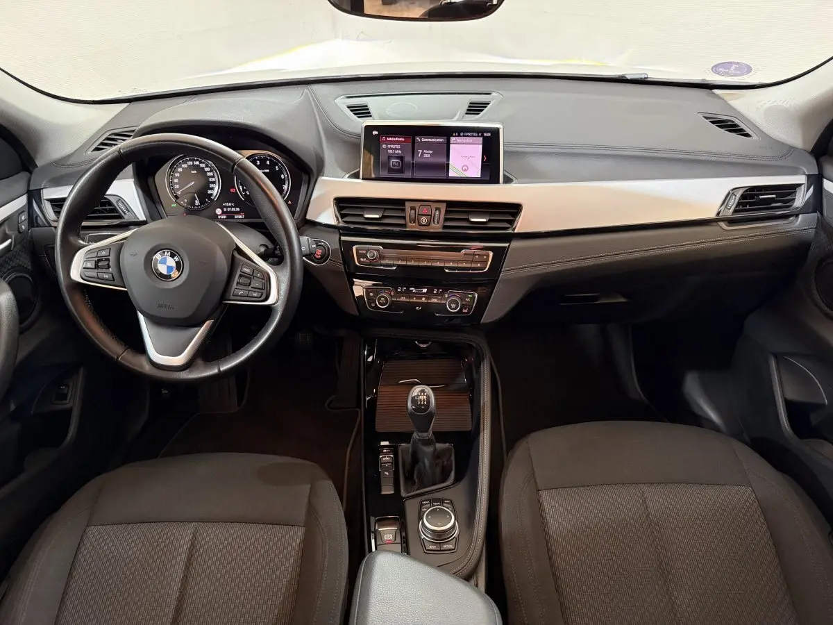 Vue intérieure avant du BMW X2 sDrive18i Lounge 2023, tableau de bord noir avec écran central et volant cuir multifonctions.