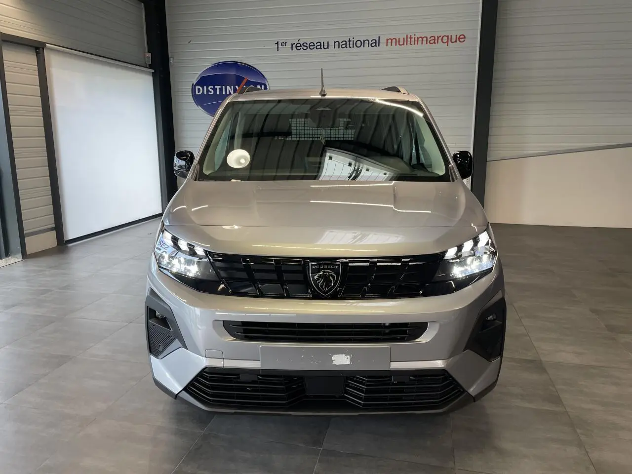 Vue frontale du Peugeot Rifter gris Artense avec calandre noire et signature lumineuse LED allumée en intérieur showroom.