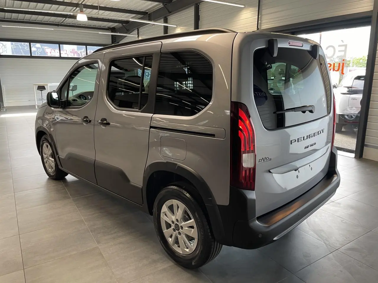 Vue 3/4 arrière droit du Peugeot Rifter gris Artense en showroom, avec portes latérales coulissantes et feux arrière allumés.