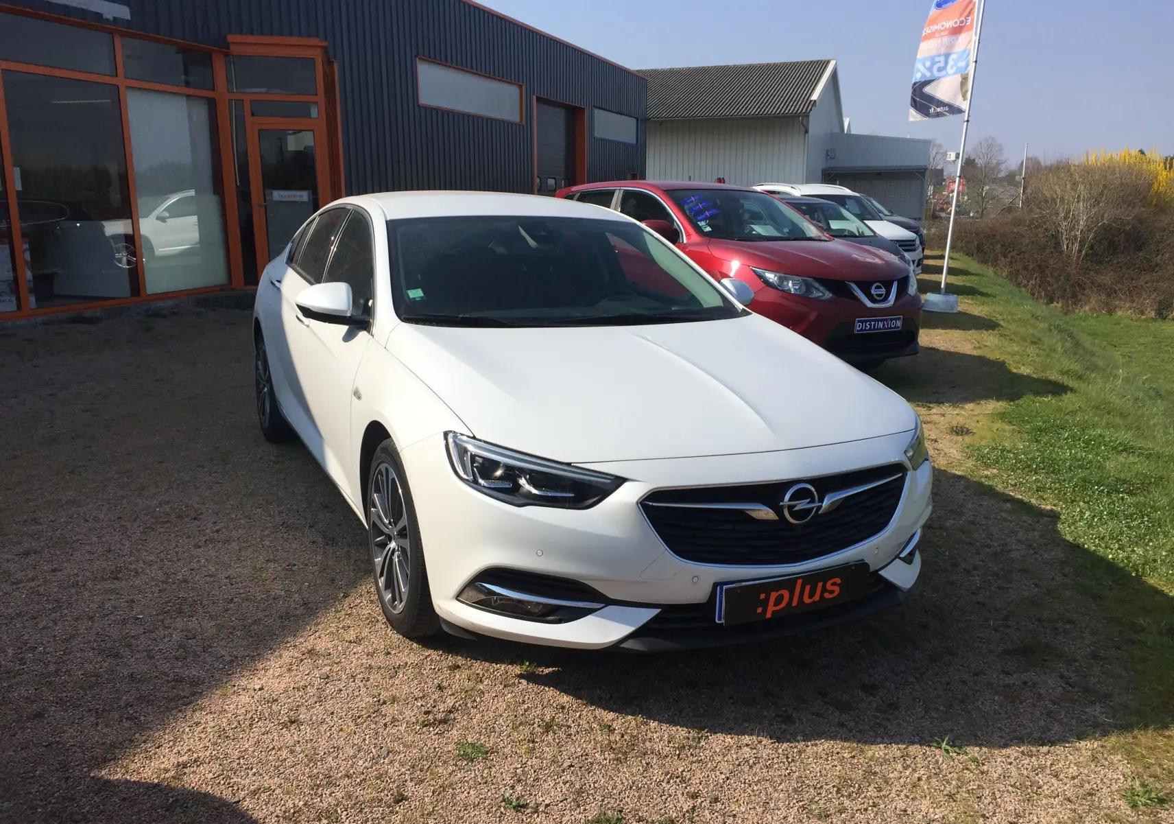 Opel Insignia Grand Sport blanc nacré vue 3/4 avant, stationnée devant un bâtiment avec d'autres voitures en arrière-plan.